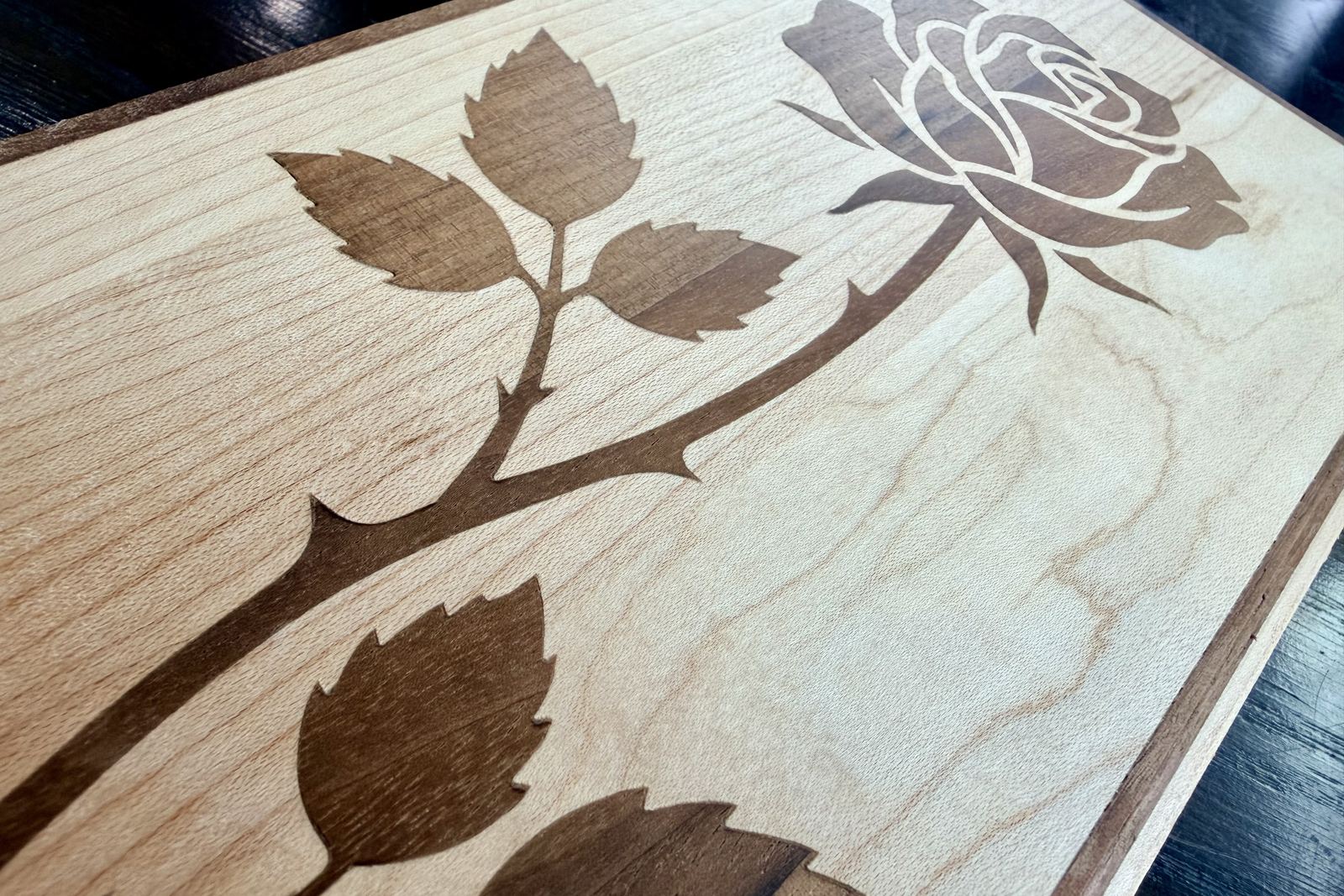 A walnut rose inlaid into a maple board, the stem and leaves seated
        flush with tight joints.